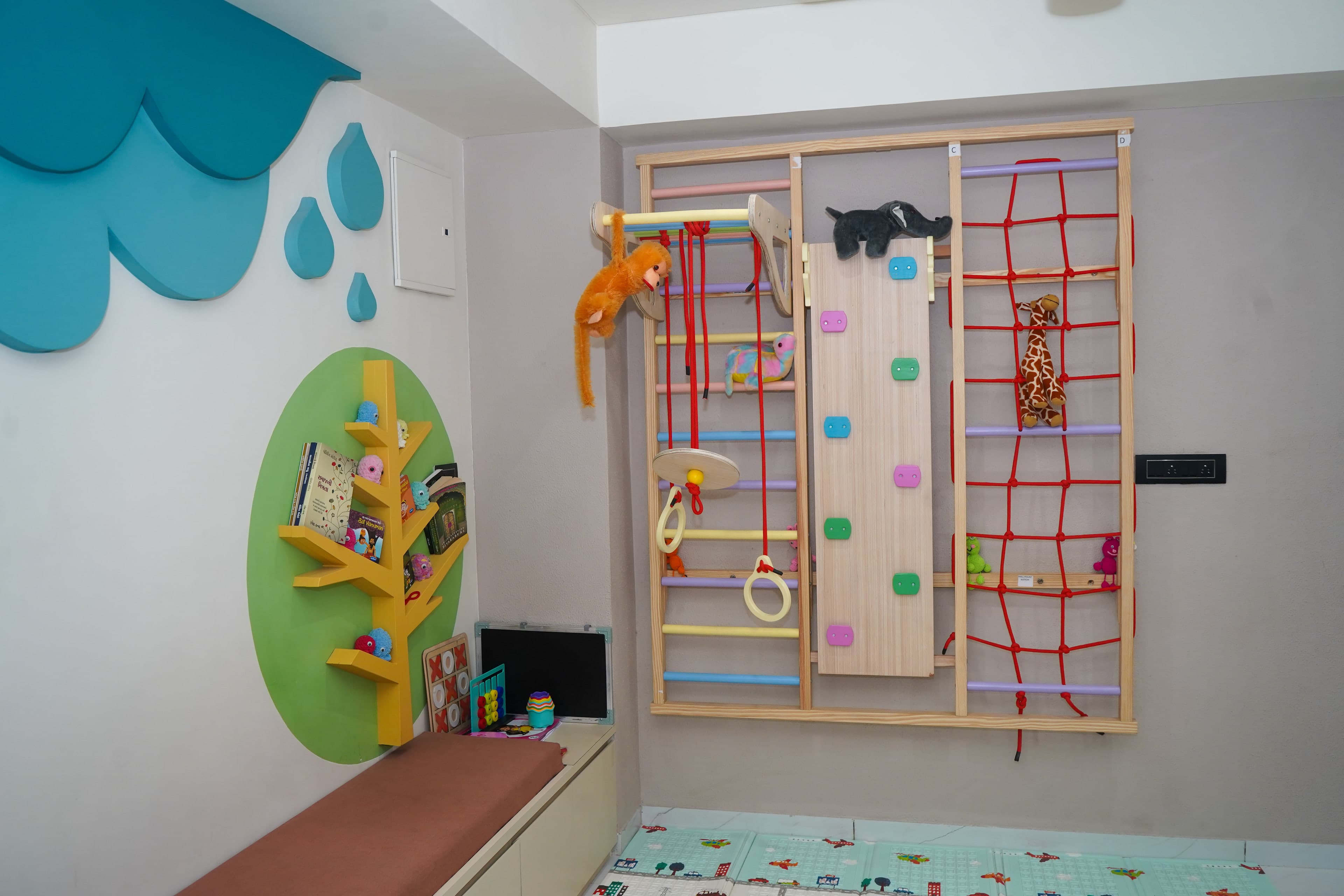 The play gym's reading side, a teal cloud-and-raindrop wall sculpture, a yellow tree bookshelf on a green disc, and bench seating with the climbing frame beyond