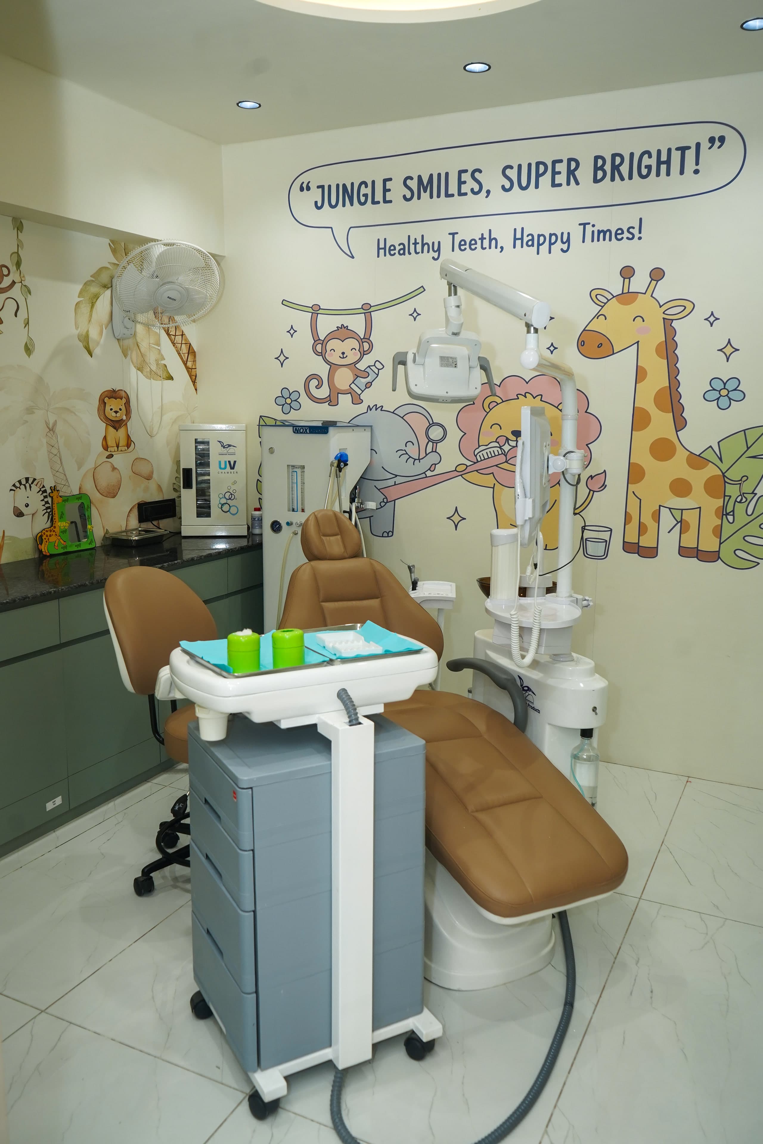 Jungle Smiles treatment room, a camel-leather dental chair beneath a 'Jungle Smiles, Super Bright!' mural of a lion brushing its teeth, an elephant with a mirror and a monkey with toothpaste, with sage cabinetry and the laughing gas unit beside the chair